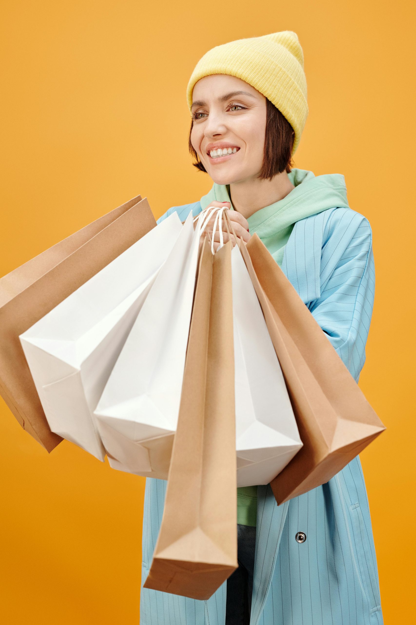 Smiling woman with paper bags on vibrant yellow background, enjoying shopping day.